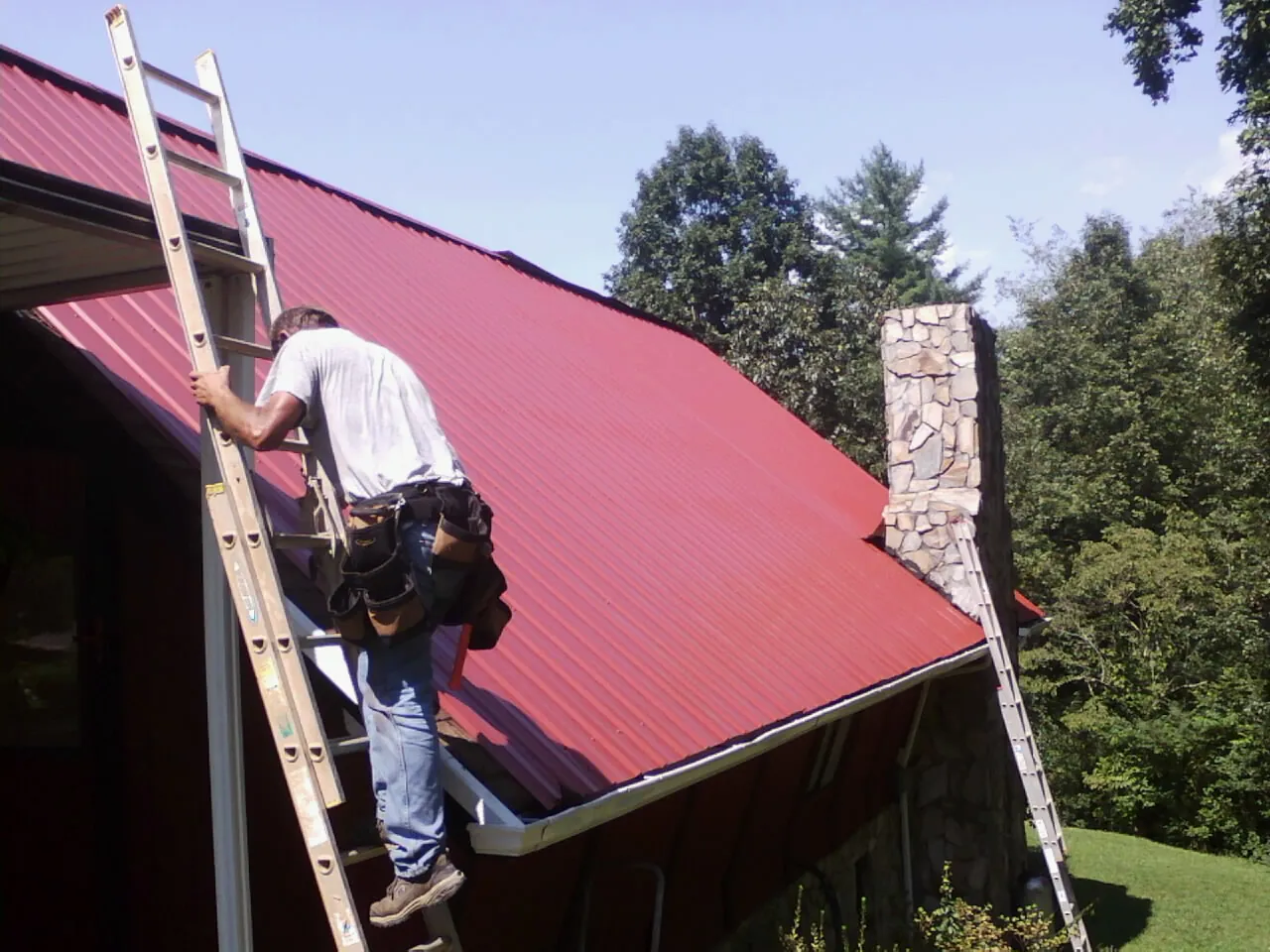 Roofer on a ladder beside a red metal roof and stone chimney — Soffit & Fascia Repair by Roofing Brothers in Bonaparte